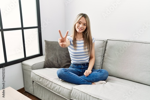 Asian young woman sitting on the sofa at home smiling looking to the camera showing fingers doing victory sign. number two.