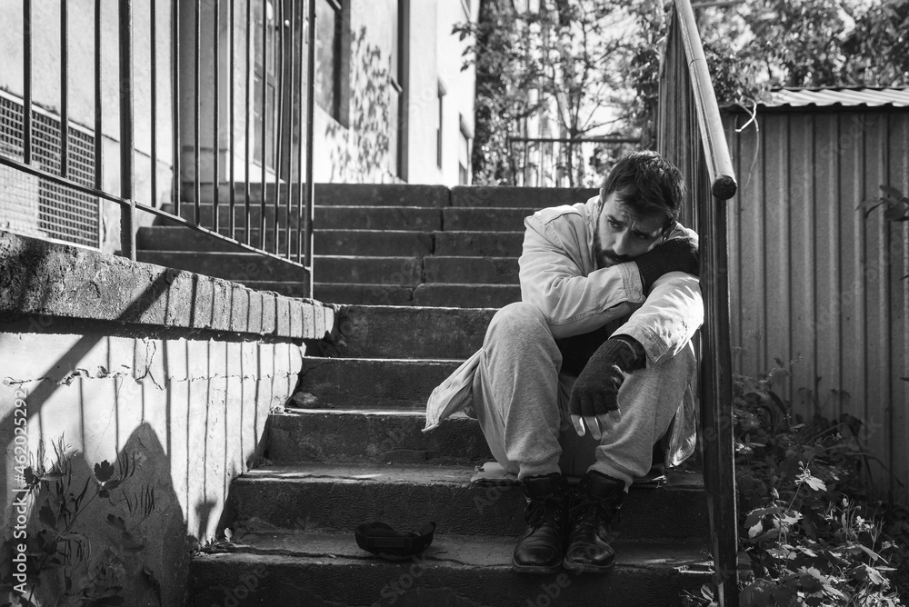 Fototapeta premium Young hungry sick and depressed homeless man sitting on the stairs of the building in the city street to rest after searching for food and to be alone out of the crowd, social documentary concept