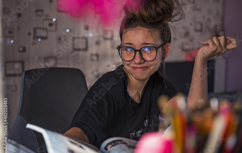 girl studying in her room at the table holding a textbook in her hand. She thinks about the information she just read. Her glasses reflect the book she is reading. photo