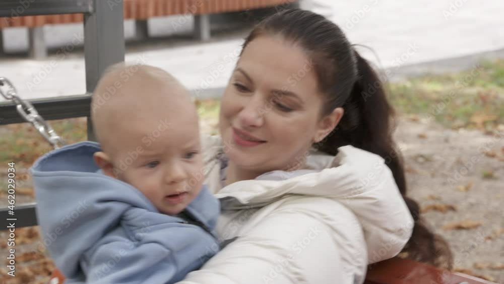 Woman with her child sitting on wooden bench swing in city park, mom enjoying the sunny autumn day with her baby outdoors. Mother with a kid. Happy family. High quality 4k footage Woman with her child sitting on wooden bench swing in city park, mom enjoying the sunny autumn day with her baby outdoors. Mother with a kid. Happy family. High quality 4k footage