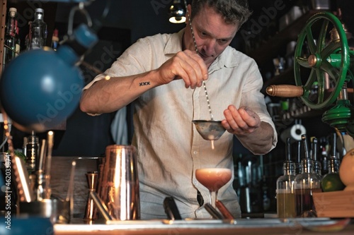 Professional bartender preparing a cocktail in a low glass using a shaker and a colander, view of a cheerful guy working in a fancy cocktail bar in night club in Lisbon, Portugal.