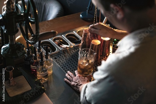 An expert bartender mixing a cocktail in a glass mixer with a long spoon  standing behind the counter of his beautiful cocktail bar in a night club.