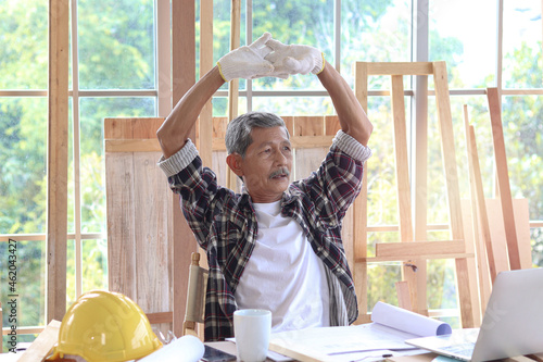 Elderly Asian carpenter with mustache taking a break and drinking a coffee, relaxing senior craftsman setting in working desk at carpentry workshop photo