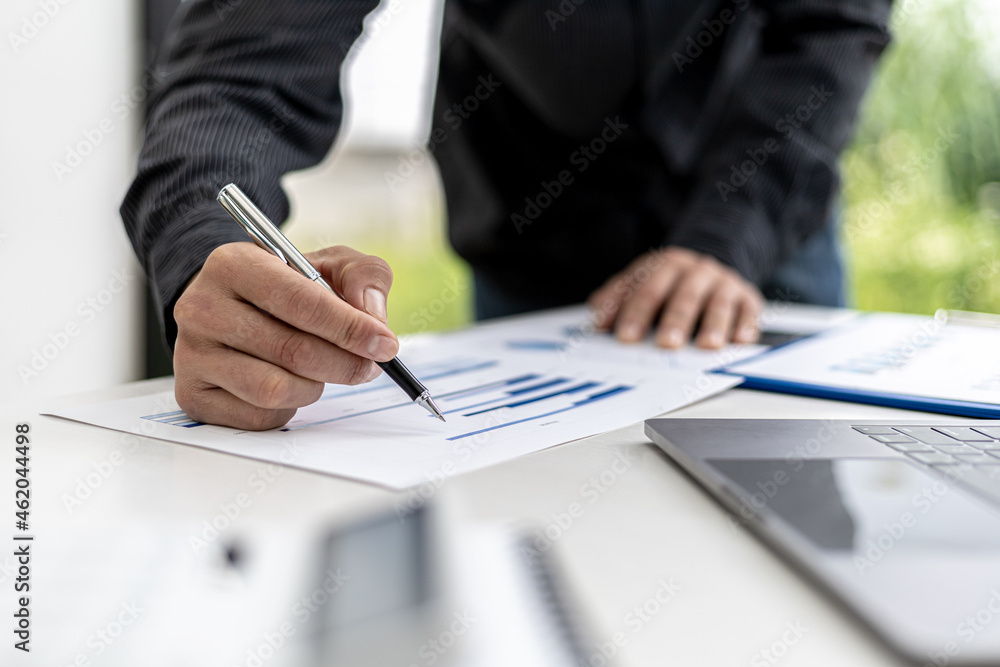 A close-up view of a businessman holding a pen pointing at a bar chart on a corporate finance document, he is meeting with senior management, with the theme of the meeting being Company Finance. A close-up view of a businessman holding a pen pointing at a bar chart on a corporate finance document, he is meeting with senior management, with the theme of the meeting being Company Finance.