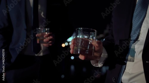 Group of unrecognizable business people cheers alcohol in transparent glasses. Adult male persons in suits spend time together at night city view close. Celebrating deal or success in business concept