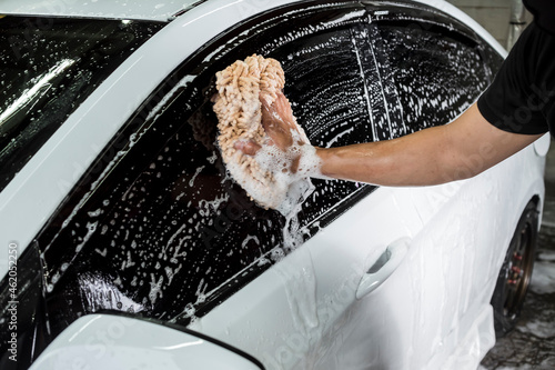 Cleaning the window of a white car with a microfiber wash mitt soaked in shampoo to lift grime and dirt. At a carwash or auto detailing shop.