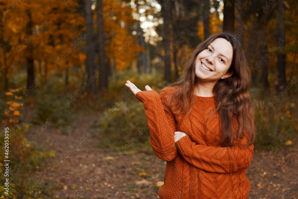 Young happy woman smiling, standing in a park against a background of an autumn forest, pointing with one hand to the side, as if demonstrating something, in a brown warm sweater, with long wavy hair. Young happy woman smiling, standing in a park against a background of an autumn forest, pointing with one hand to the side, as if demonstrating something, in a brown warm sweater, with long wavy hair.
