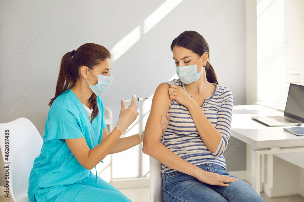 Highly qualified nurse gives Covid-19 vaccine to responsible female patient at modern medical center. Beautiful young woman wearing mouth covering getting flu shot during seasonal vaccination campaign - obrazy, fototapety, plakaty 