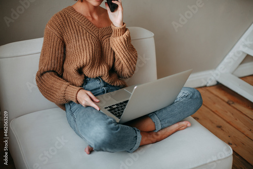 Young brunette girl with short hair in a brown sweater and blue jeans sitting on a white armchair and talking on a cell phone and holds a laptop in his hands