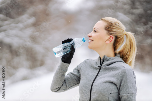 Obraz na plátne Happy sportswoman standing in nature at snowy winter day, taking a break and drinking fresh water