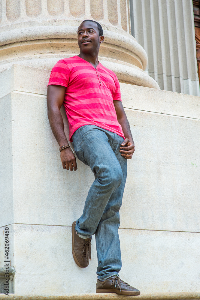 Wearing a red, pink stripe Henley V Neck T shirt, gray pants, leather sneakers, a young black guy is standing against the base of a column, bending over a leg, waiting for you.. Wearing a red, pink stripe Henley V Neck T shirt, gray pants, leather sneakers, a young black guy is standing against the base of a column, bending over a leg, waiting for you..