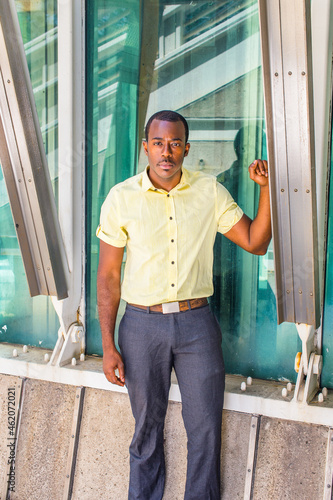 Dressing in a light yellow shirt, gray pants, a young attractive black guy is standing by a triangle structure, looking at you..