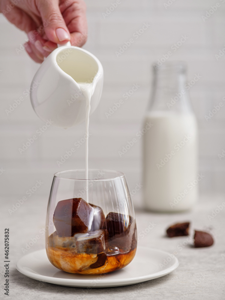 Pouring milk into a glass of espresso coffee with frozen coffee cubes on bright background.