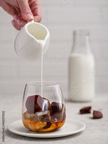 Pouring milk into a glass of espresso coffee with frozen coffee cubes on bright background.