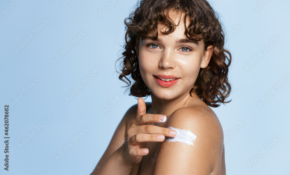 Body and care. Beautiful young woman apply cream on her shoulder, moisturizing with skincare product, smiling tender at camera, blue background