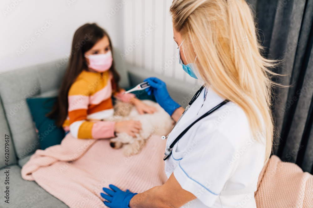 Sick little girl covered in blanket is lying on couch while doctor checks girl's body temperature using thermometer. Sick little girl covered in blanket is lying on couch while doctor checks girl's body temperature using thermometer.