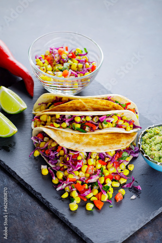 Vegetarian tacos with various vegetables, guacamole and sliced lime on dark background. Tacos with sweet corn, purple cabbage and pepper on a slate board. Copy space