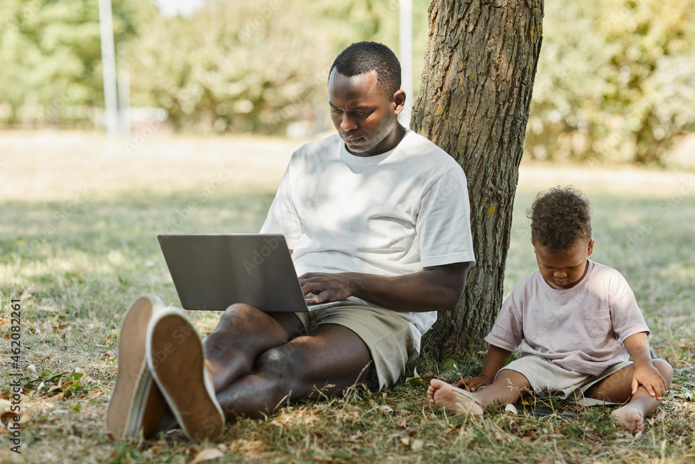 Full length portrait of young African-American man using laptop outdoors in park with cute baby son Full length portrait of young African-American man using laptop outdoors in park with cute baby son