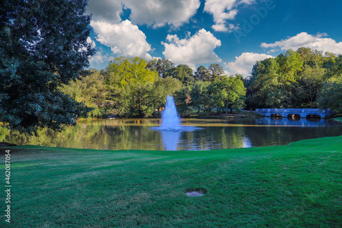 a gorgeous shot of the silky green lake water with a fountain in the middle of the lake with a stone bridge surrounded by lush green and autumn colored trees at Lenox Park in Brookhaven Georgia USA photo