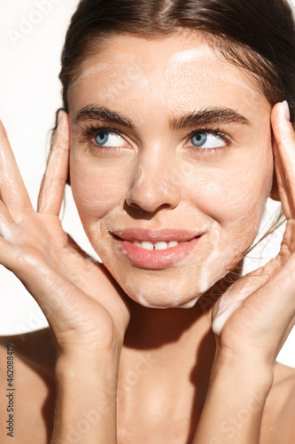 Vertical portrait of young woman washing her face with cleansing foam gel, smiling happy, cleaning her facial skin, standing over white background