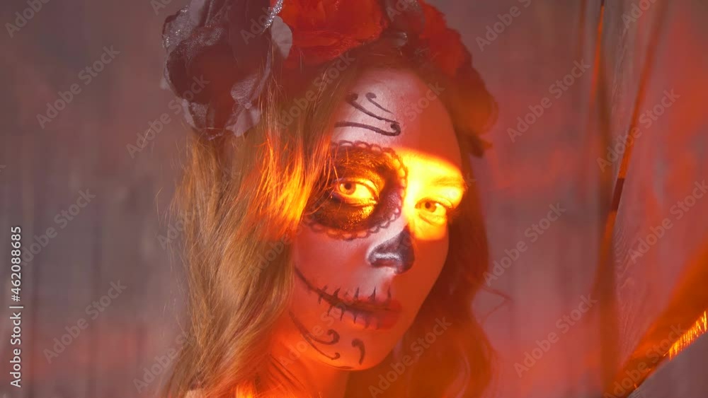 A mysterious portrait of a woman with Halloween makeup and roses on her head in red light and smoke turns and looks into the camera. Mysticism, abstraction and witchcraft. A mysterious portrait of a woman with Halloween makeup and roses on her head in red light and smoke turns and looks into the camera. Mysticism, abstraction and witchcraft.