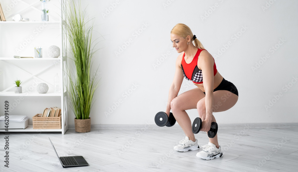 Power gym. Athletic woman. Online training. Strong body. Smiling sportive lady in sportswear doing deep squats with dumbbells in hands looking laptop in light room interior copy space.