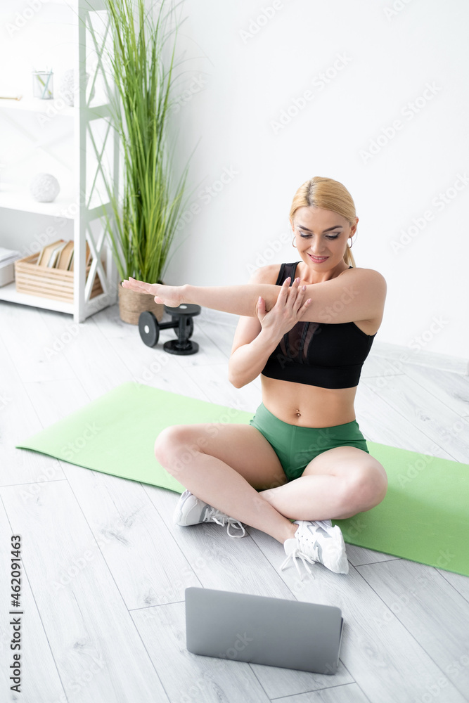 Home fitness. Happy woman. Enjoying aerobic. Healthy lifestyle. Athletic lady in sportswear doing warming stretching exercise hands looking laptop in light room interior. Home fitness. Happy woman. Enjoying aerobic. Healthy lifestyle. Athletic lady in sportswear doing warming stretching exercise hands looking laptop in light room interior.