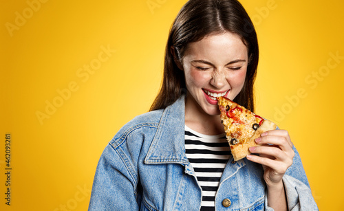 Smiling girl eating pizza, biting slice and looking aside satisfied, trying tasty food, concept of takeout order in pizzeria, eating-out in restaurants, yellow background