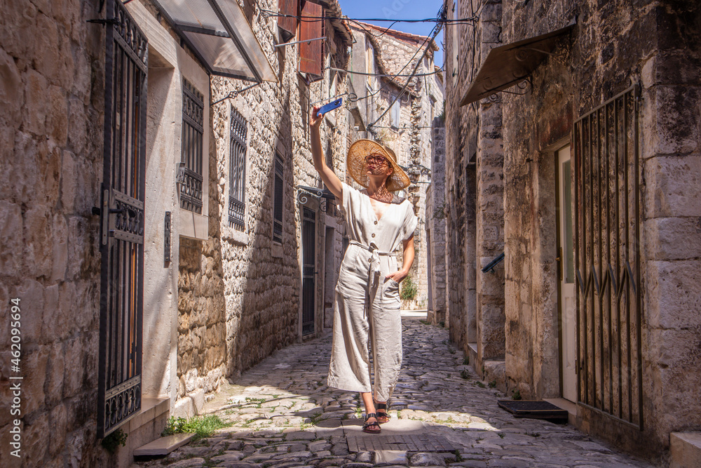 Beautiful blonde young female traveler wearing straw sun hat, taking selfie while sightseeing and enjoying summer vacation in an old traditional costal town at Adriatic cost, Croatia. Beautiful blonde young female traveler wearing straw sun hat, taking selfie while sightseeing and enjoying summer vacation in an old traditional costal town at Adriatic cost, Croatia.