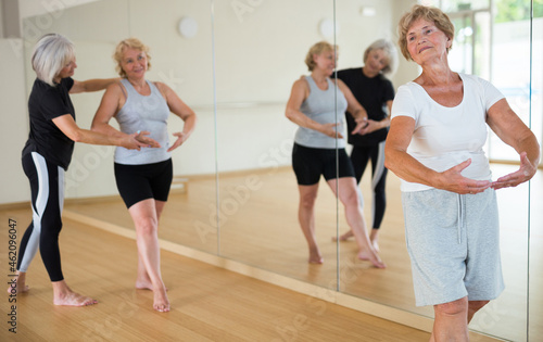 Positive elderly woman practicing ballet dance moves during group class in choreographic studio.