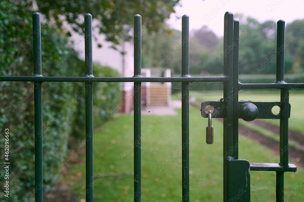 Padlocked closed green metal gate with out of focus park and lawn in background Padlocked closed green metal gate with out of focus park and lawn in background