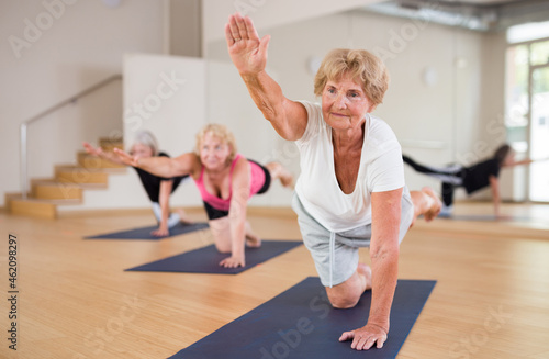 Mature active women who attend group classes in a fitness studio perform a stretching exercise