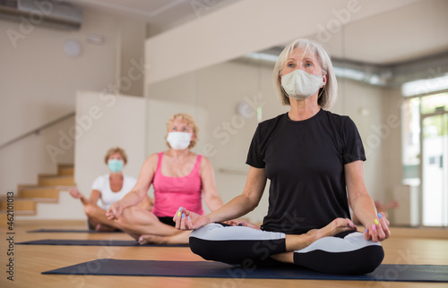 Sporty aged woman in protective face mask sitting on mat in lotus positions during group yoga training. Precautions in coronavirus pandemic.