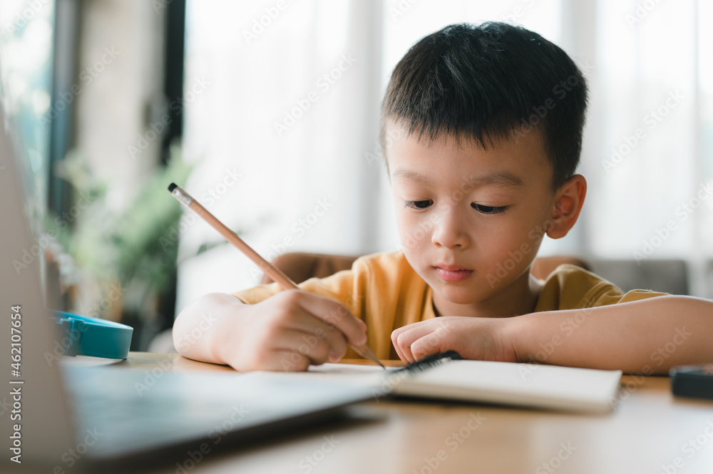 Asian Boy Doing homework with the intention. Child boy holding pencil writing, studying at home on a laptop to study from home during Covid-19 pandemic lockdown. Online education, Homeschooling. Asian Boy Doing homework with the intention. Child boy holding pencil writing, studying at home on a laptop to study from home during Covid-19 pandemic lockdown. Online education, Homeschooling.