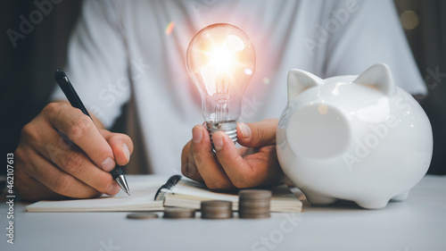 saving energy and money concept. idea for save or investment. businessman holding lightbulb on piggy bank and coins stacking on desk with note for planning money finance. photo