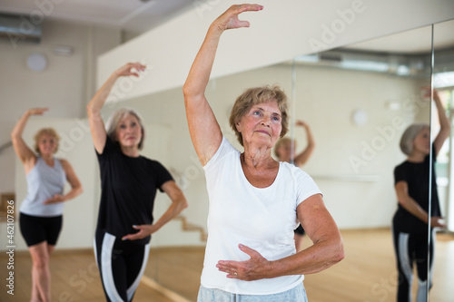 Positive elderly woman practicing ballet dance moves during group class in choreographic studio.
