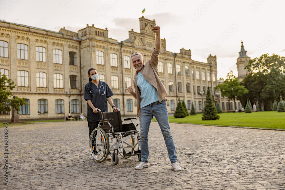 Obraz premium Joyful disabled handicapped mature man can walk again. Recovering patient standing up from his wheelchair, young nurse assisting him outdoors