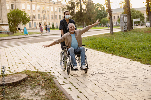 Excited mature disabled man in wheelchair wearing headphones having fun during a walk in the city assisted by young nurse in protective face mask