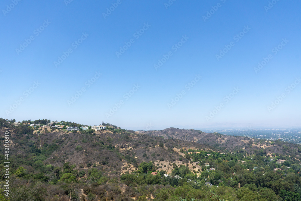 View of Hollywood Hills seen from Mulholland Drive on a sunny summer day. Houses, trees and dry patches of land stretching across the valley