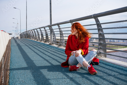 a young adult red-haired woman in a red jacket  blue jeans and red sneakers is sitting on the sidewalk. In the background there is a metal fence  sky and lampposts. Daylight  everyday lifestyle.