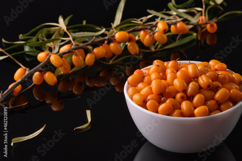 A sea buckthorn berry in a white cup stands on a black reflective surface in close-up. Nearby lies a branch of sea buckthorn.