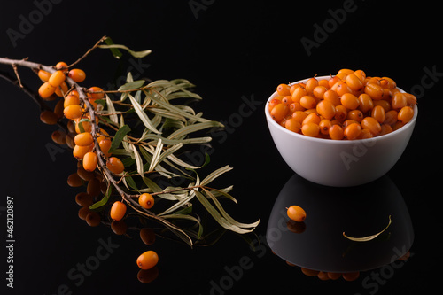 a branch of sea buckthorn and a white cup filled with sea buckthorn berries on a black reflective surface