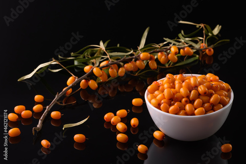 sea buckthorn berry in a white cup stands on a black reflective surface. A branch lies nearby and sea buckthorn berries are scattered.