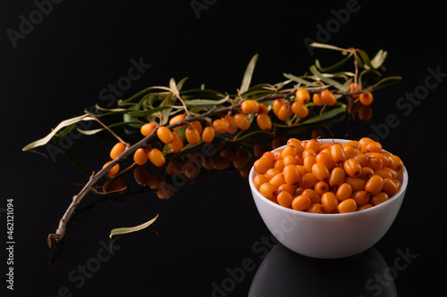 Sea buckthorn berry in a white cup stands on a black reflective surface. Nearby lies a branch of sea buckthorn.