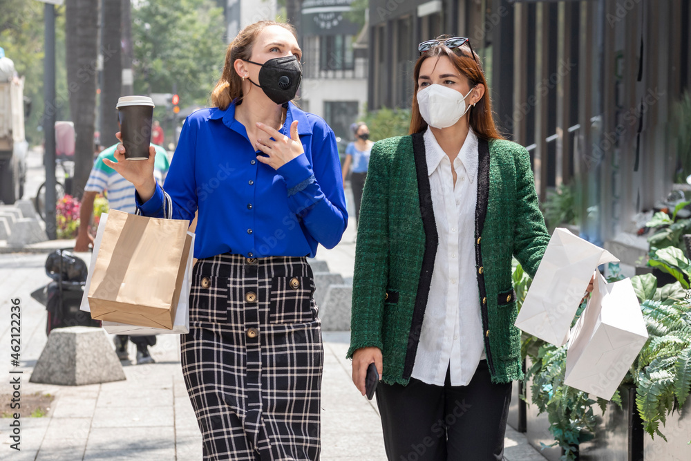 Two young women are walking down the street wearing face masks talking happily after shopping carrying bags and a glass of coffee