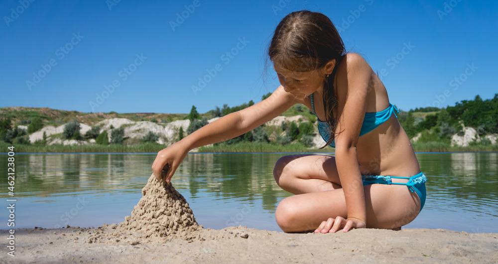 A little cute girl in a swimsuit is building a sand castle on the shore ...