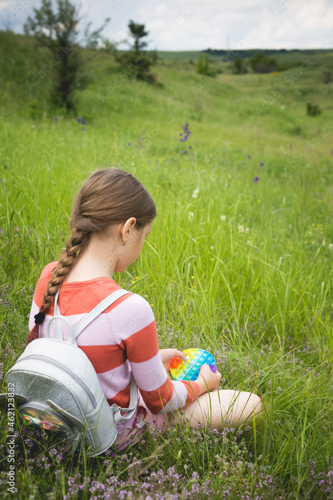 A little cute girl is sitting on the grass with a pop it - antistress toy. A little cute girl is sitting on the grass with a pop it - antistress toy.