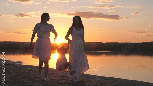 cheerful girls with small child are walking along the summer beach at sunset, happy family, raising a child together, playing jumping, childhood dream of flying, traveling on weekends in the sunshine