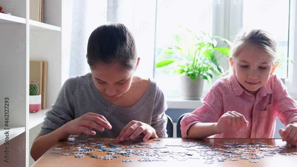 Two cute little sisters-girls are happy to make jigsaw puzzles, sitting at a table in the room. The concept of childhood, games and leisure. Two cute little sisters-girls are happy to make jigsaw puzzles, sitting at a table in the room. The concept of childhood, games and leisure.