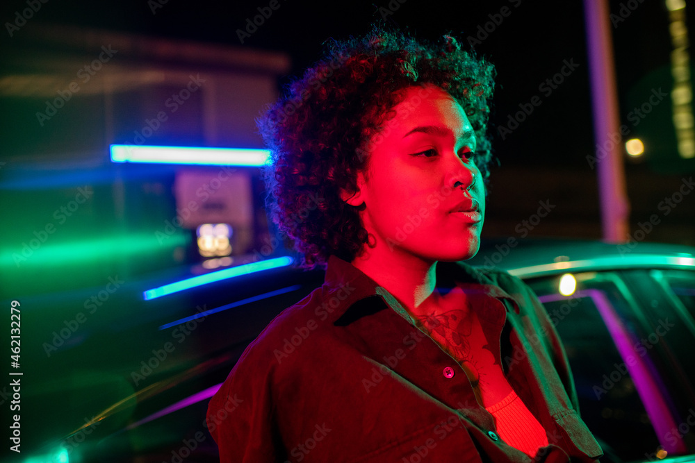 Young serene woman leaning against car in urban environment at night - obrazy, fototapety, plakaty 